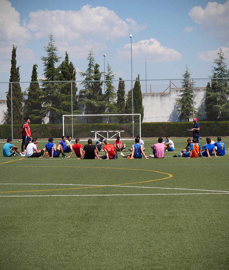 Estudiantes de la Facultad de CIencias del Deporte en una práctica de fútbol. Los estudiantes sentados en corro en el centro del campo reciben instrucciones por parte de su profesor.